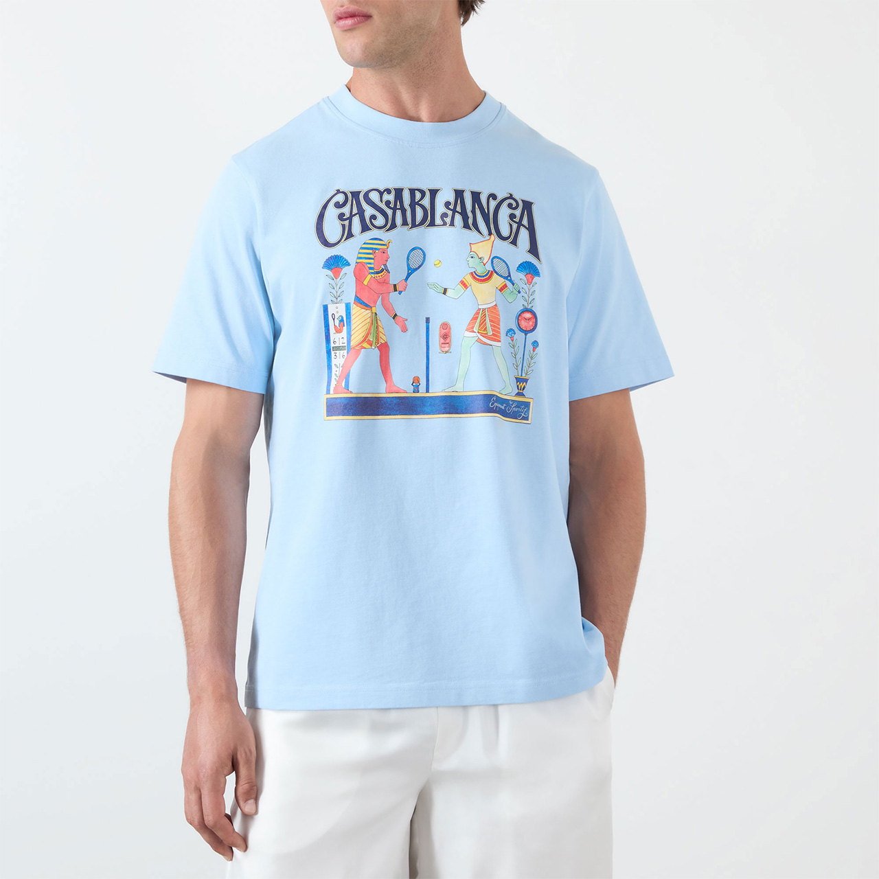 A man wearing a light blue T-shirt with a colourful illustration of two people playing tennis and the text 'CASABLANCA' above. The T-shirt is paired with white trousers.