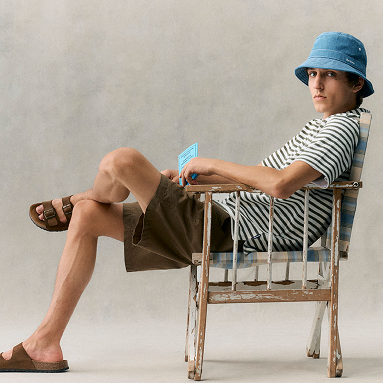 Young man wearing blue bucket hat, striped shirt, and olive shorts, relaxing on wooden deck chair