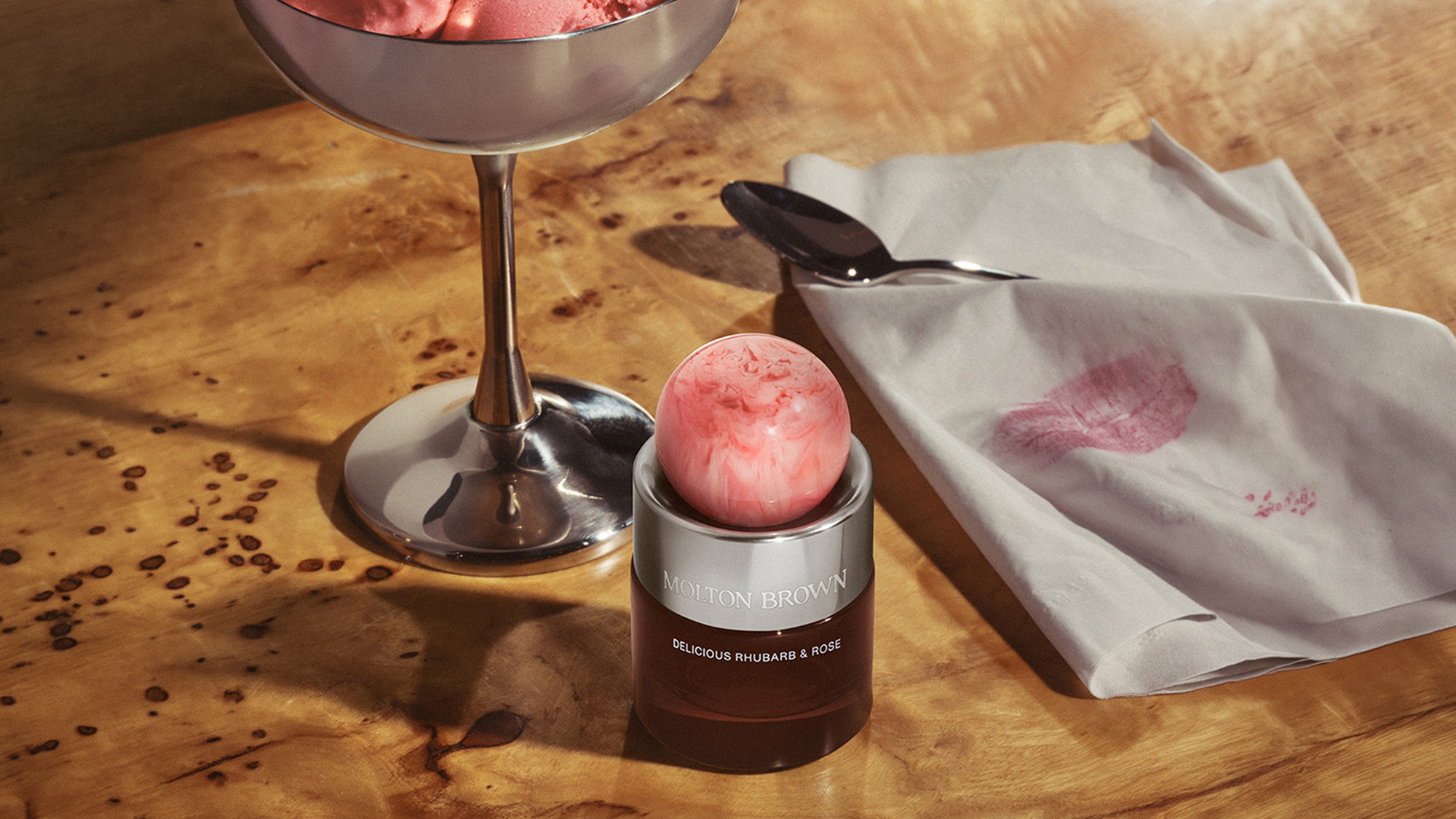 A scoop of pink ice cream sits on a silver stand, with a jar of Molton Brown Delicious Rhubarb & Rose beneath it. Nearby, a metal spoon and a napkin with a pink lipstick stain rest on a wooden surface.