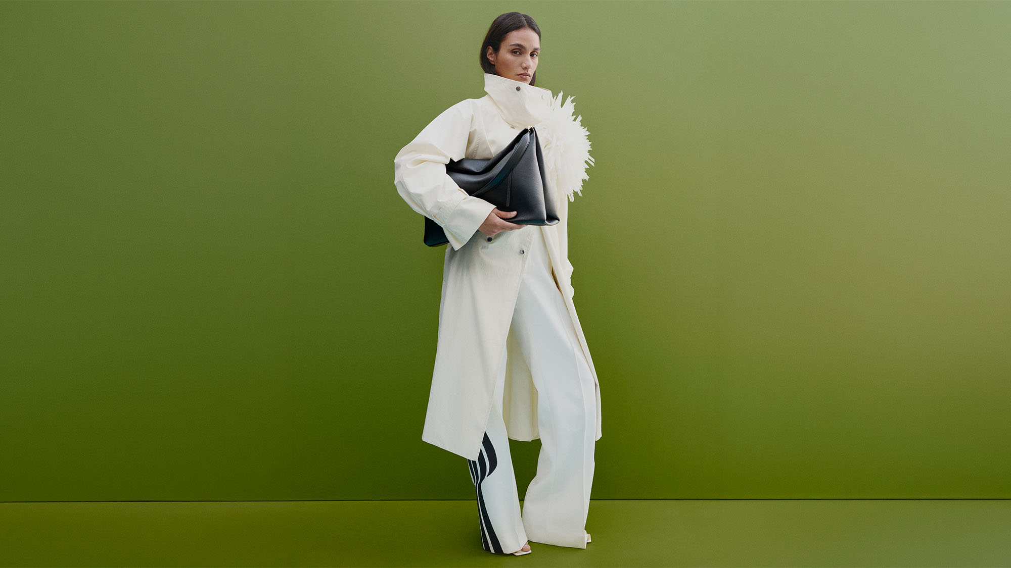 A person in a white coat and trousers poses against a green background, holding a black bag.