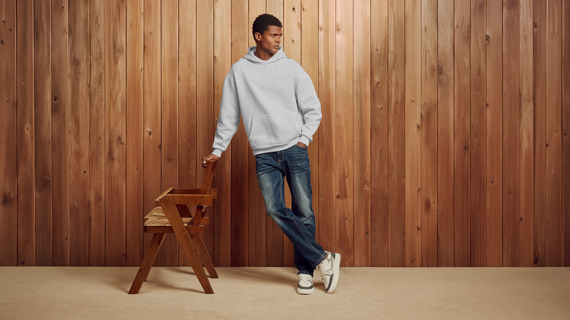 Man wearing light grey hoodie and dark blue jeans with cream trainers, standing against wooden wall backdrop