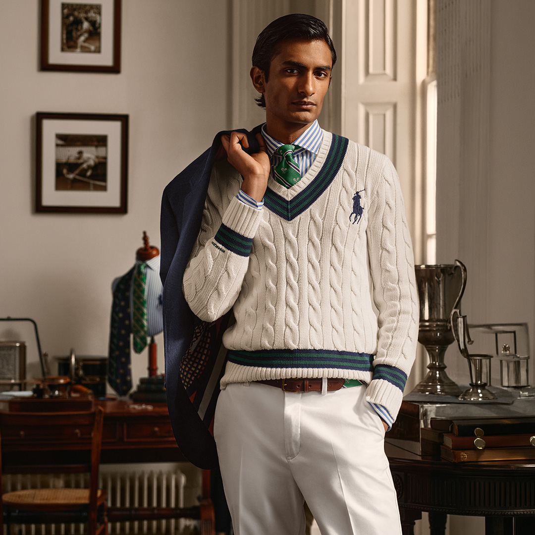 Man wearing cream cable-knit cricket jumper with green and navy stripes, white trousers, and matching tie in elegant interior setting