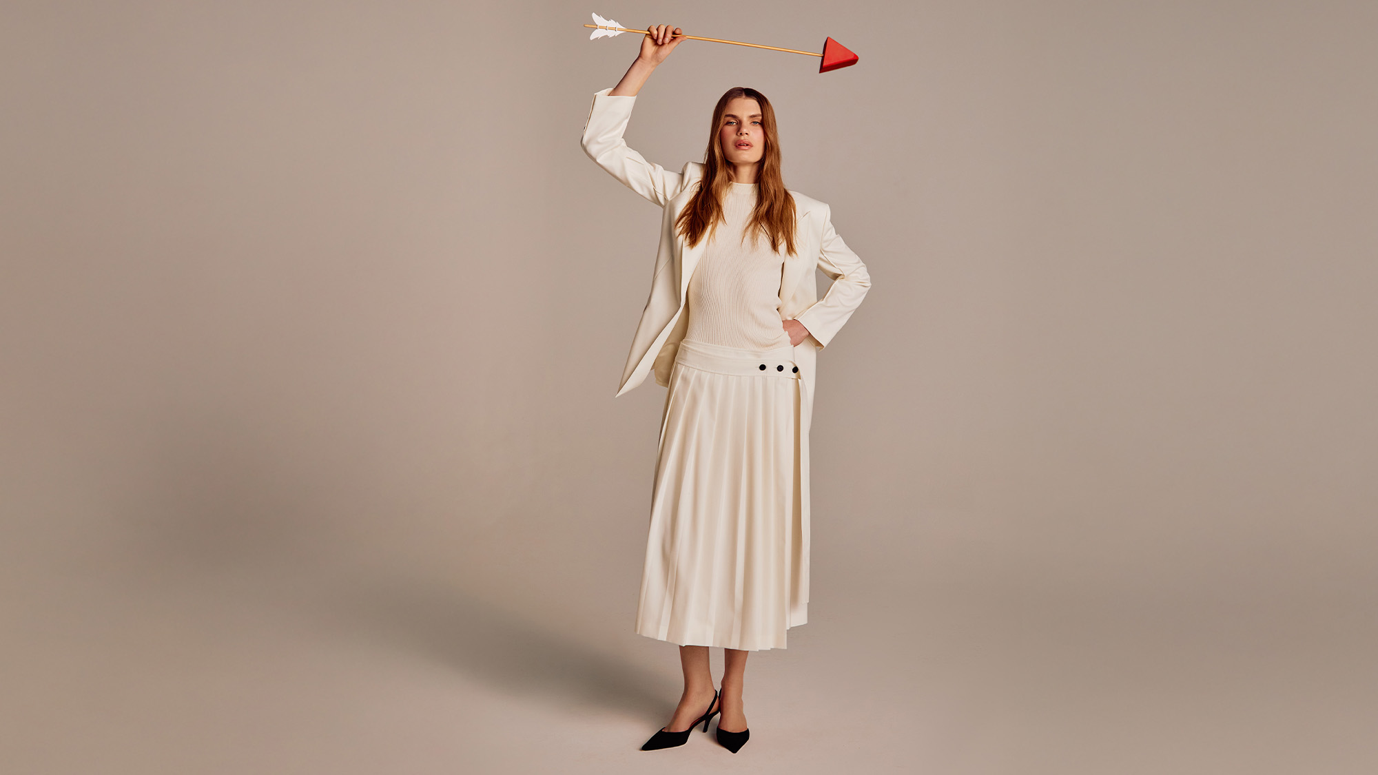 A woman in a white outfit holds a large arrow overhead, standing confidently against a neutral background.