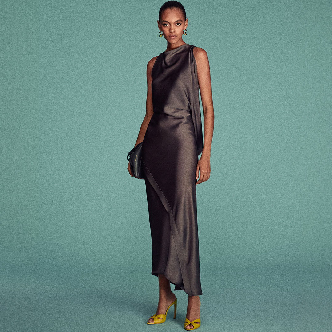 Woman in charcoal sleeveless draped midi dress with yellow heeled sandals against turquoise background