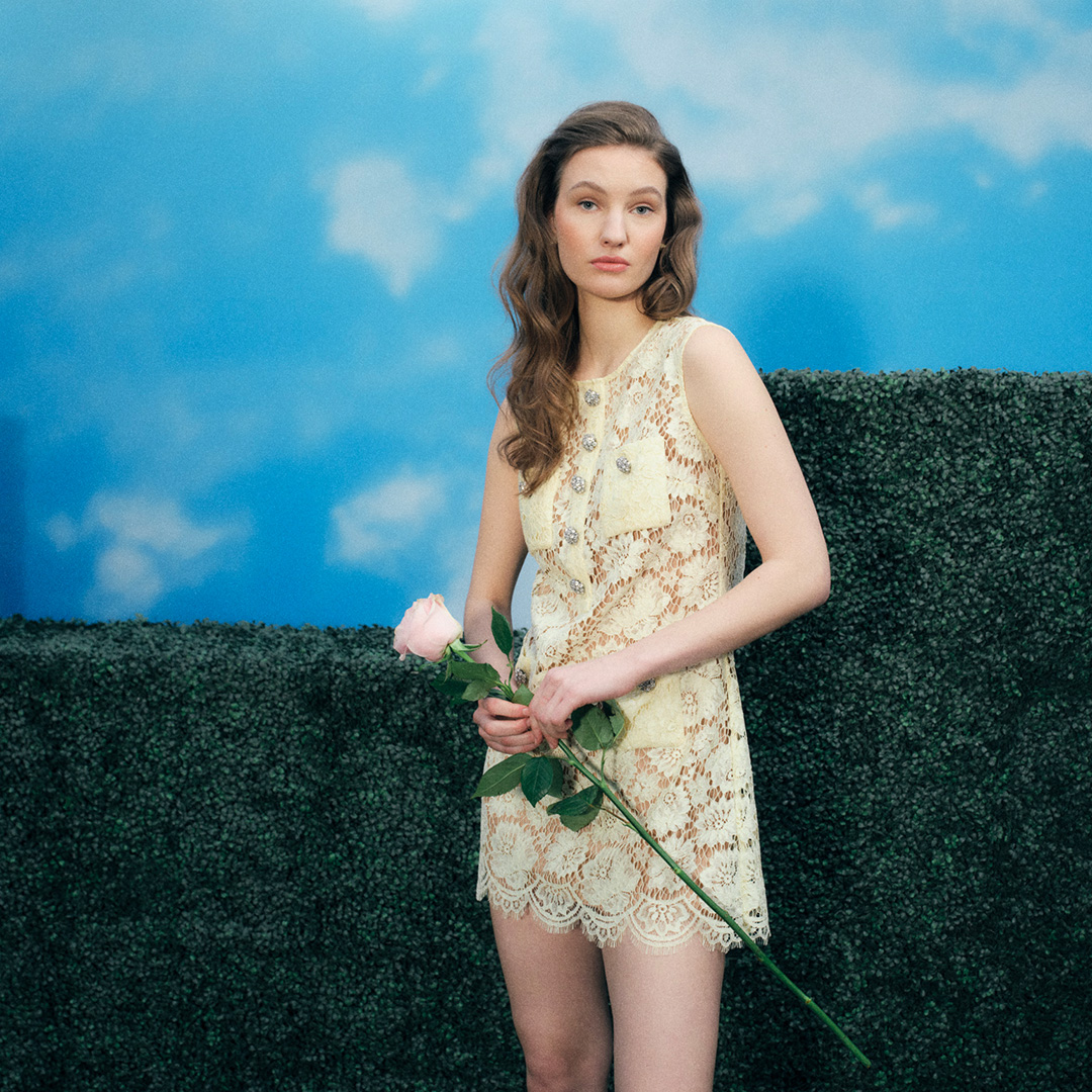 Woman wearing cream lace dress holding white rose, standing against topiary hedge backdrop