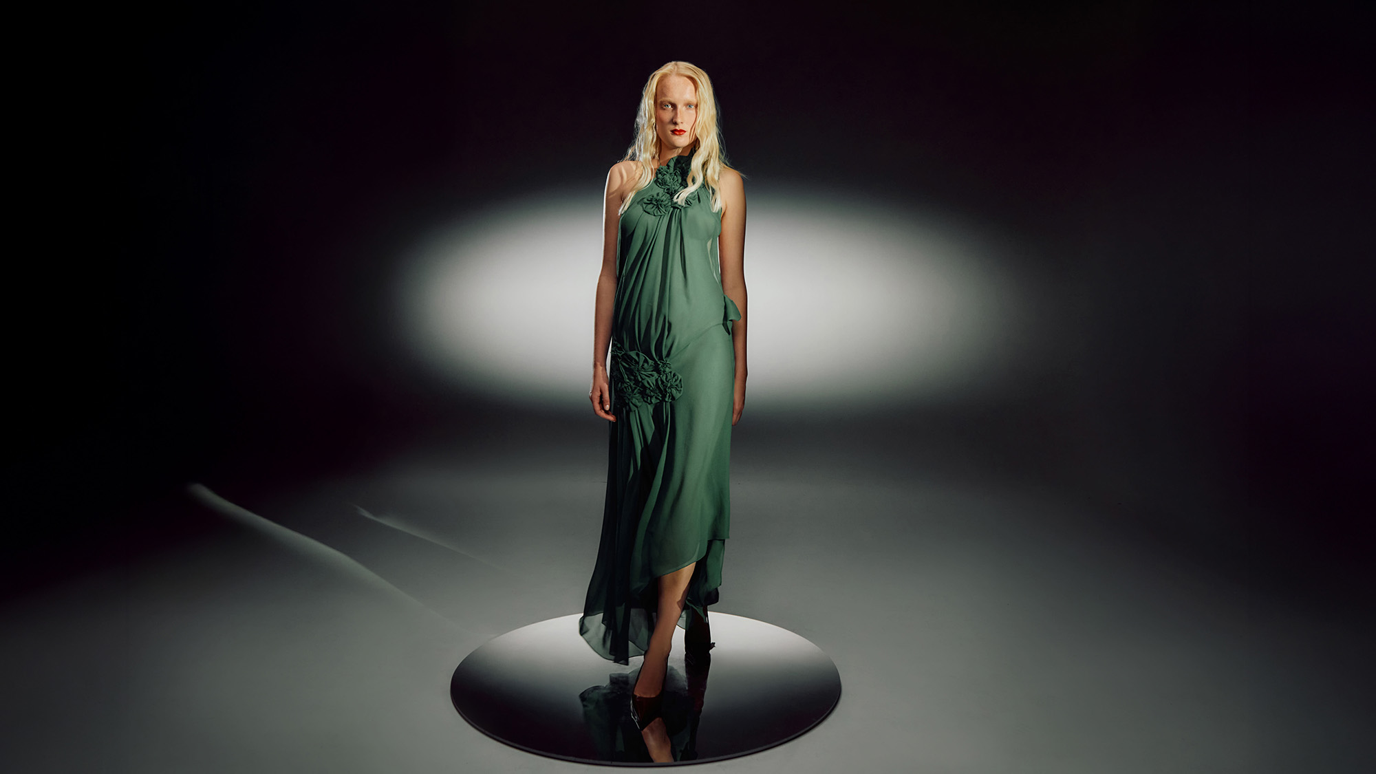 Woman wearing flowing green halterneck dress with embellished detail against dramatic lighting