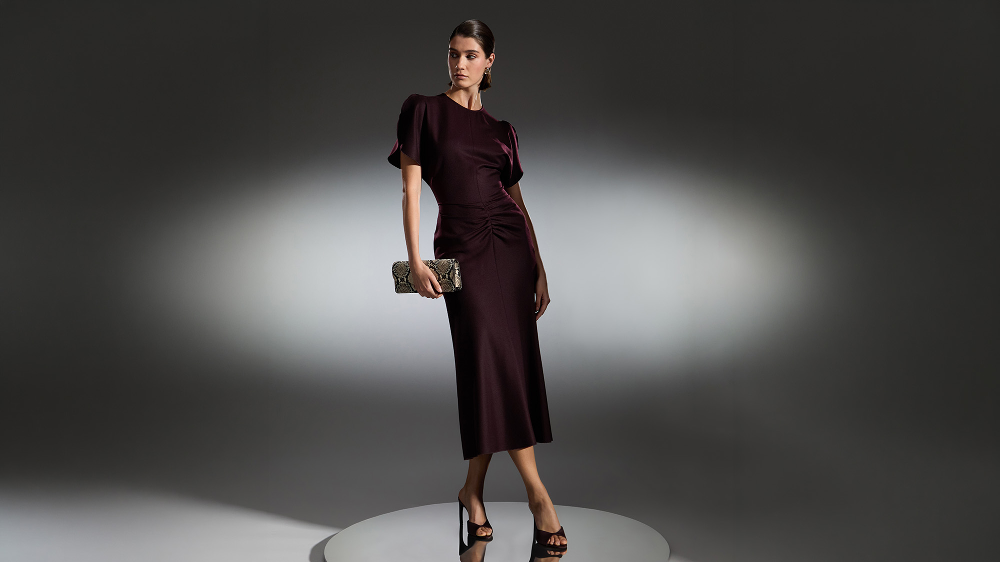 Woman wearing burgundy ruched midi dress with black heeled sandals and metallic clutch bag