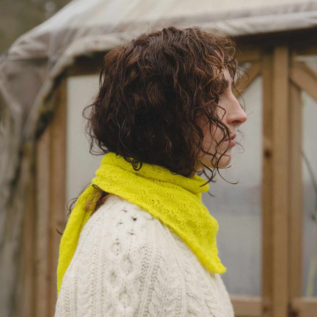 Woman with curly brown hair wearing a bright yellow scarf and cream cable-knit sweater, photographed in profile outdoors.