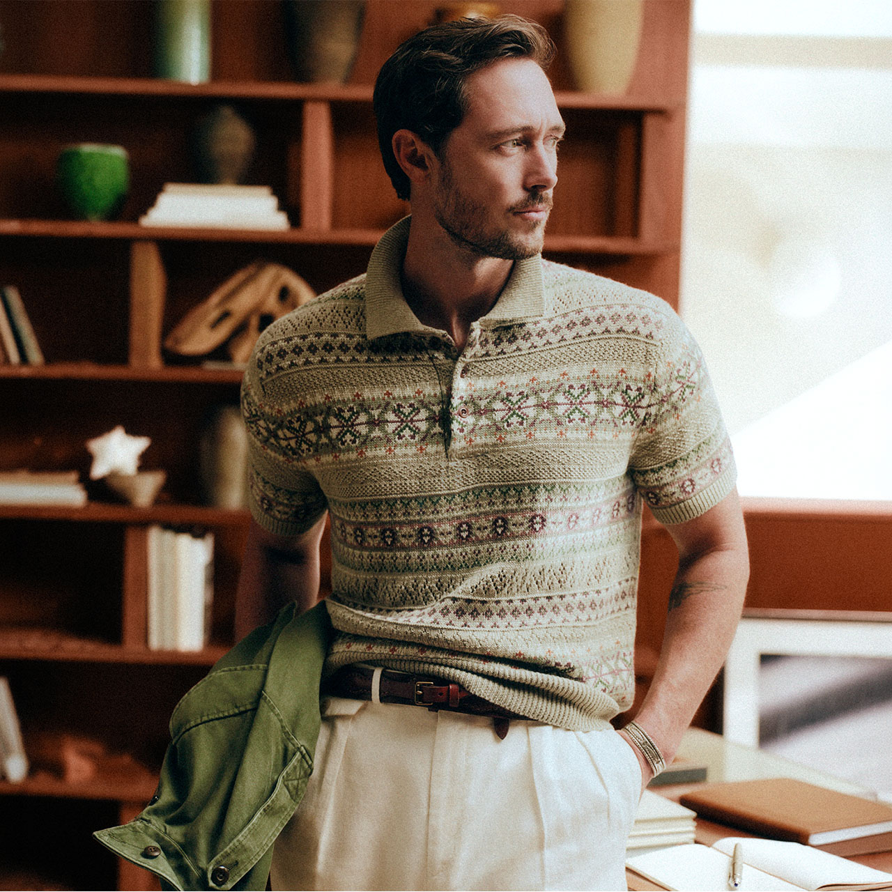 Man wearing patterned polo shirt and cream trousers, holding green jacket, standing in study with wooden shelving