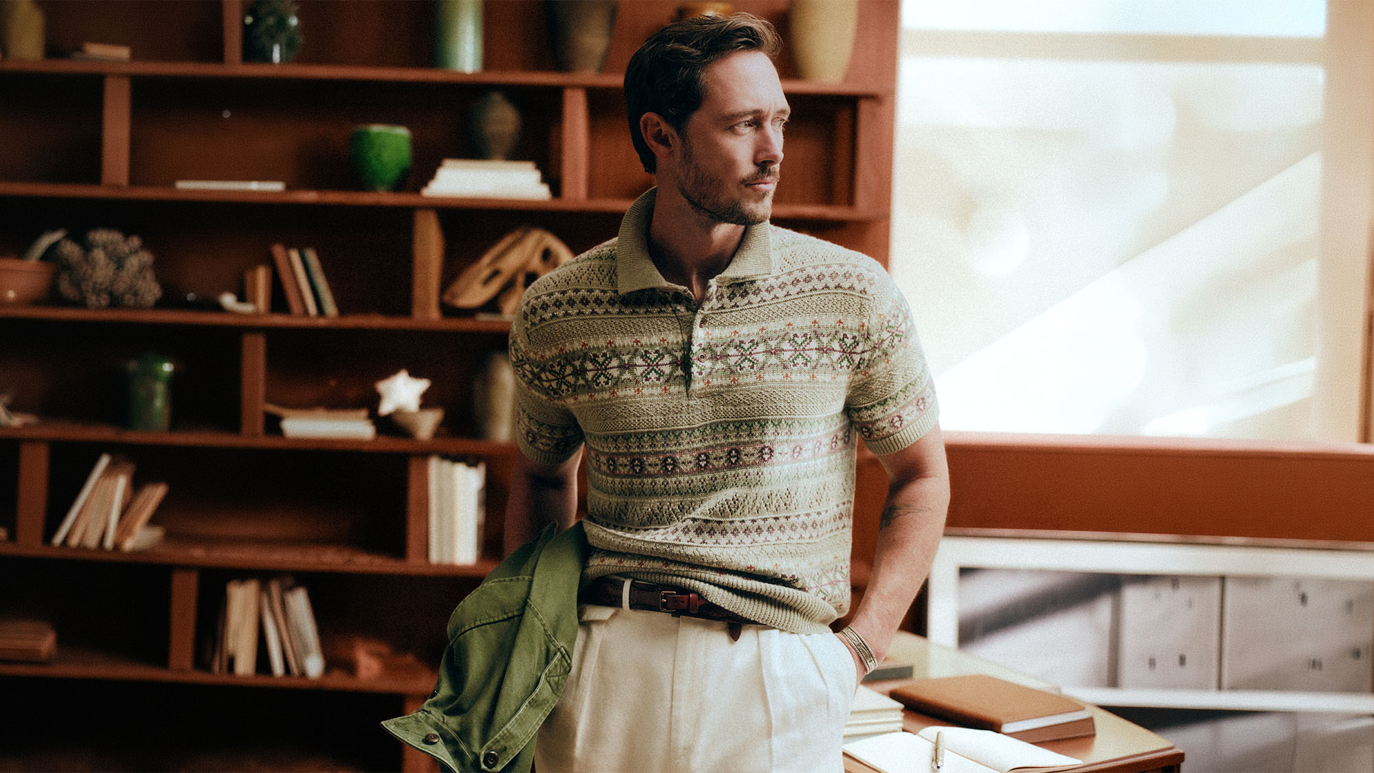 Man wearing patterned polo shirt and cream trousers, holding green jacket, standing in study with wooden shelving