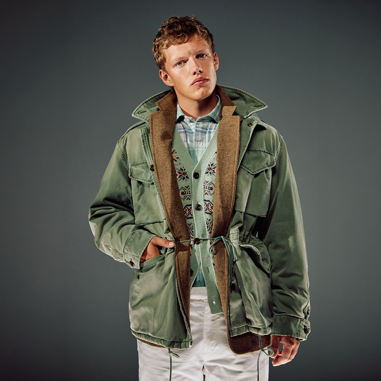 Young man wearing layered outfit with sage green jacket, brown gilet, and checked shirt over white base layer