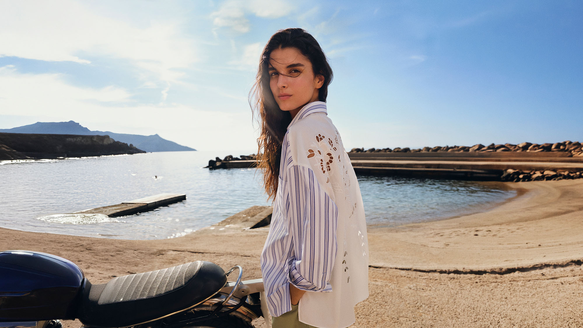 Woman on beach wearing white perforated shirt and striped blue shirt, coastal landscape background