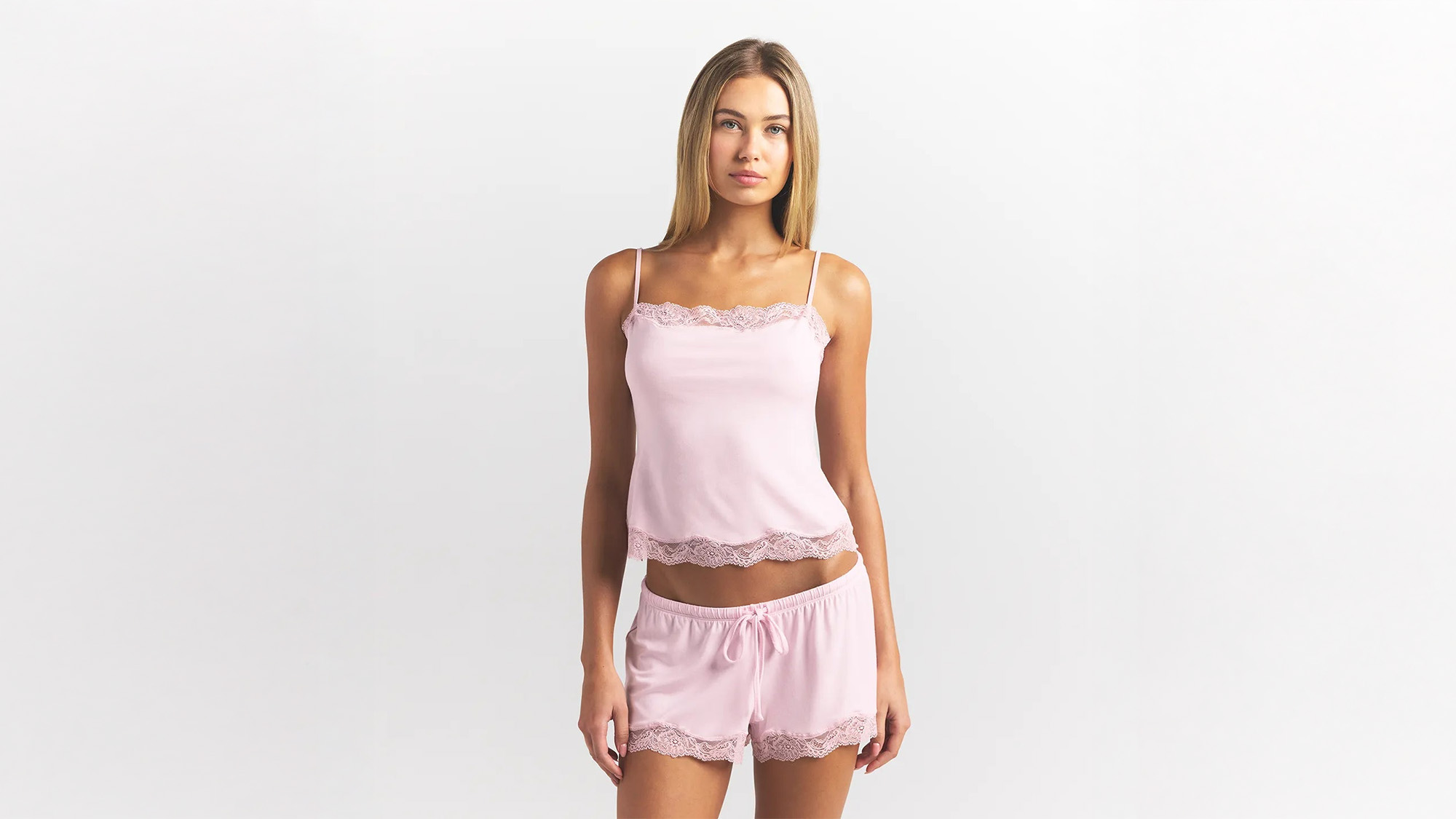 A person wearing a pink lace-trimmed camisole and shorts set, standing against a plain white background.
