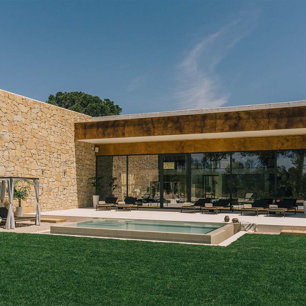 Modern house with a stone facade, large windows, and a swimming pool on a grassy yard under a clear blue sky.