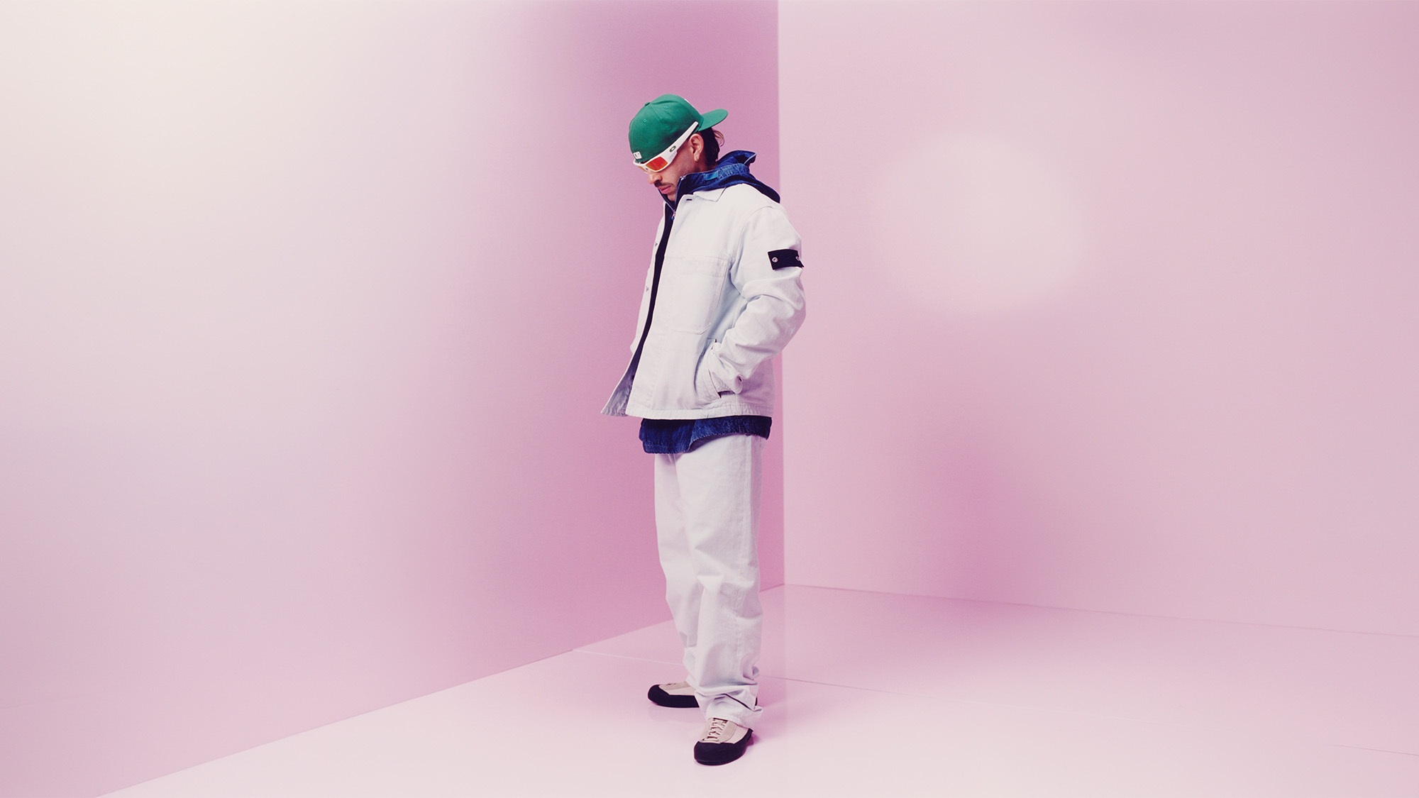 A person in a green cap, sunglasses, and layered white clothing stands in a pink room.