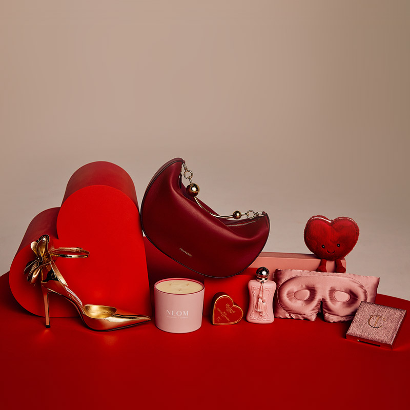 Assorted luxury items, including a gold high heel, red handbag, candle, and perfume, arranged on a red surface with heart shapes.