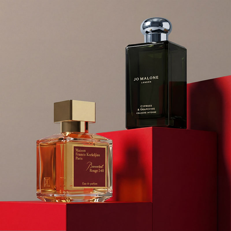 Two perfume bottles, one rectangular with a gold cap and transparent amber liquid, and one tall with a silver cap, set against bold red steps.