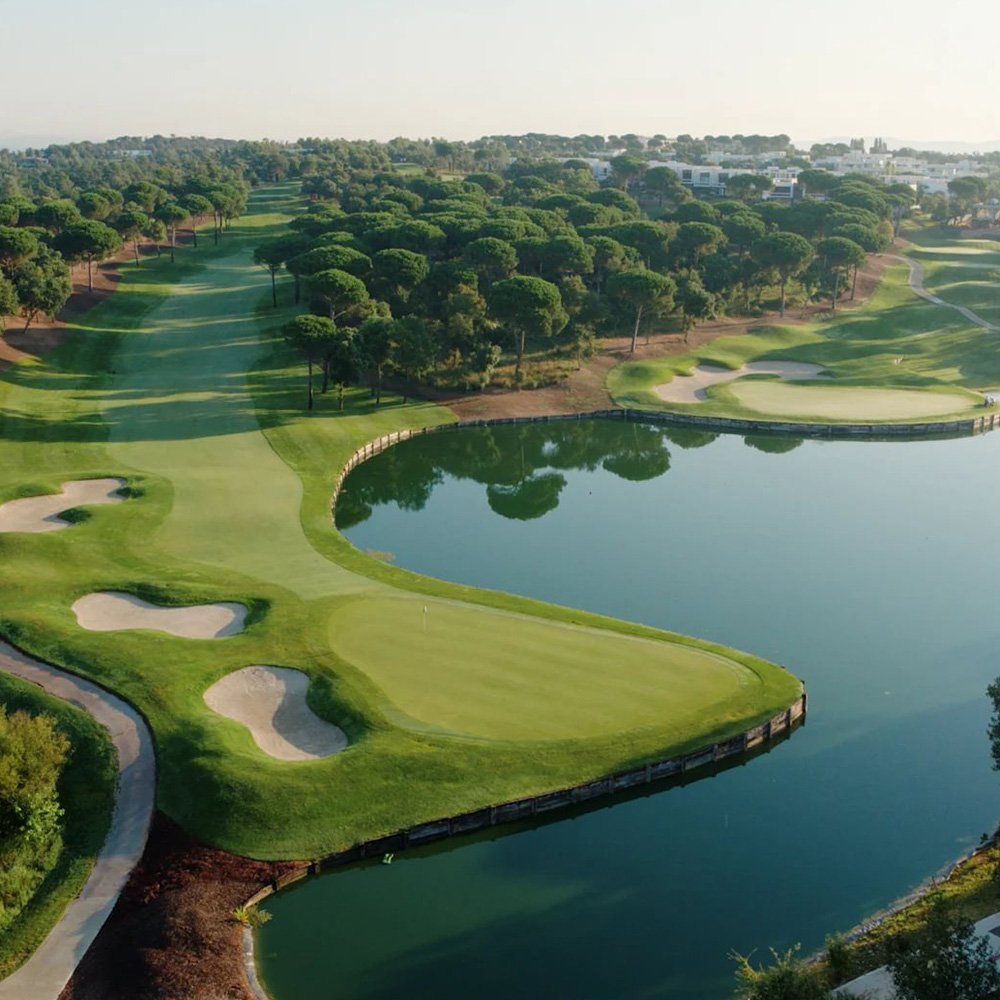 Aerial view of a lush golf course featuring greens, sand bunkers, and a reflecting pond, surrounded by dense trees.




