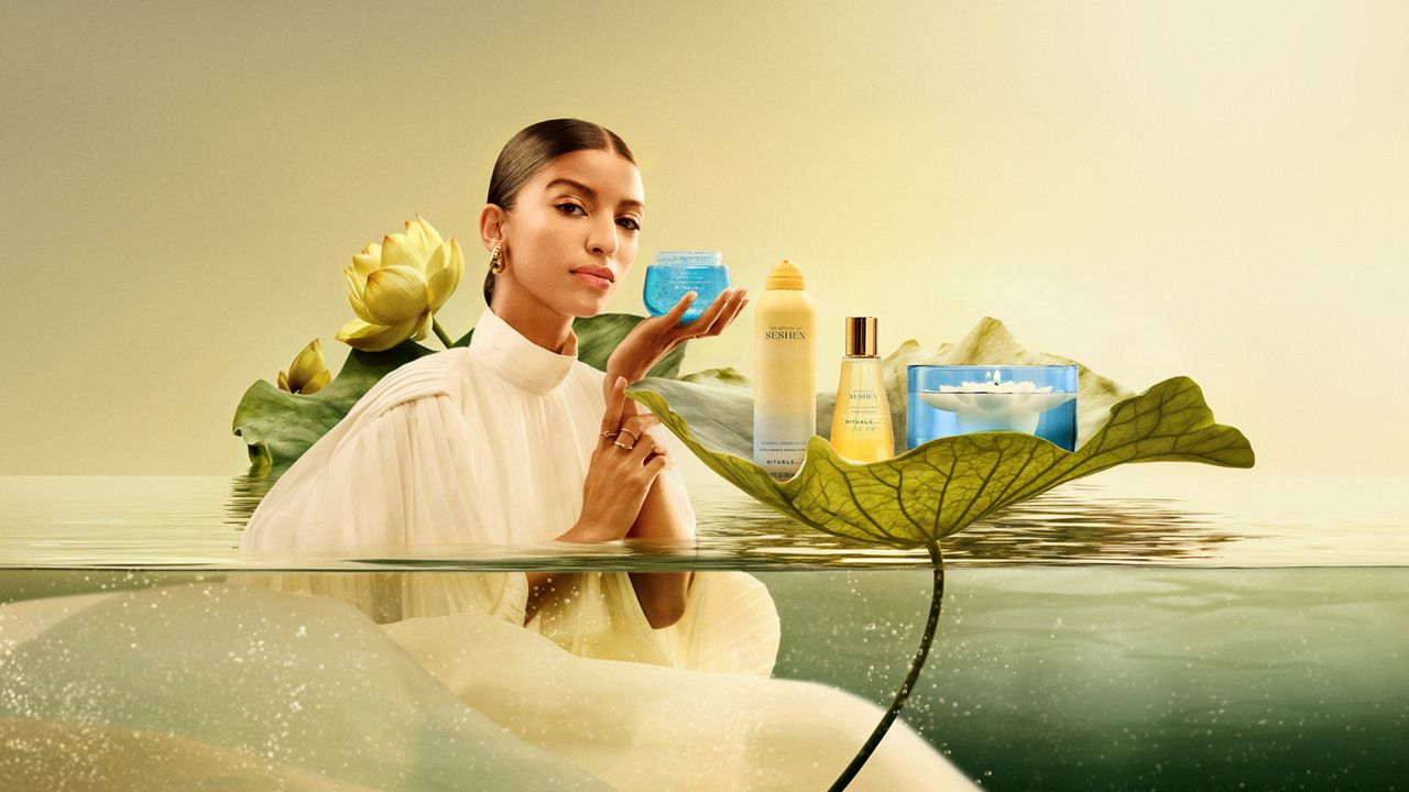 A woman in a white dress stands in water, holding a blue jar. Skincare products are displayed on a large leaf.