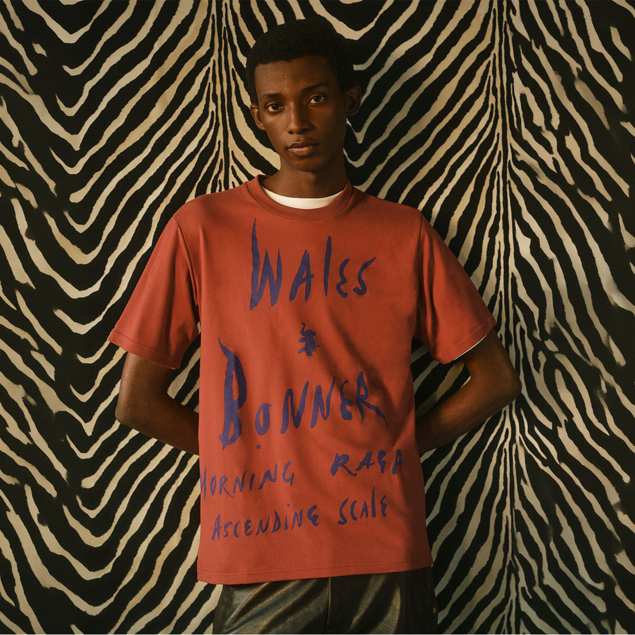 Man wearing rust-coloured t-shirt with blue text graphic, standing against black and cream striped background