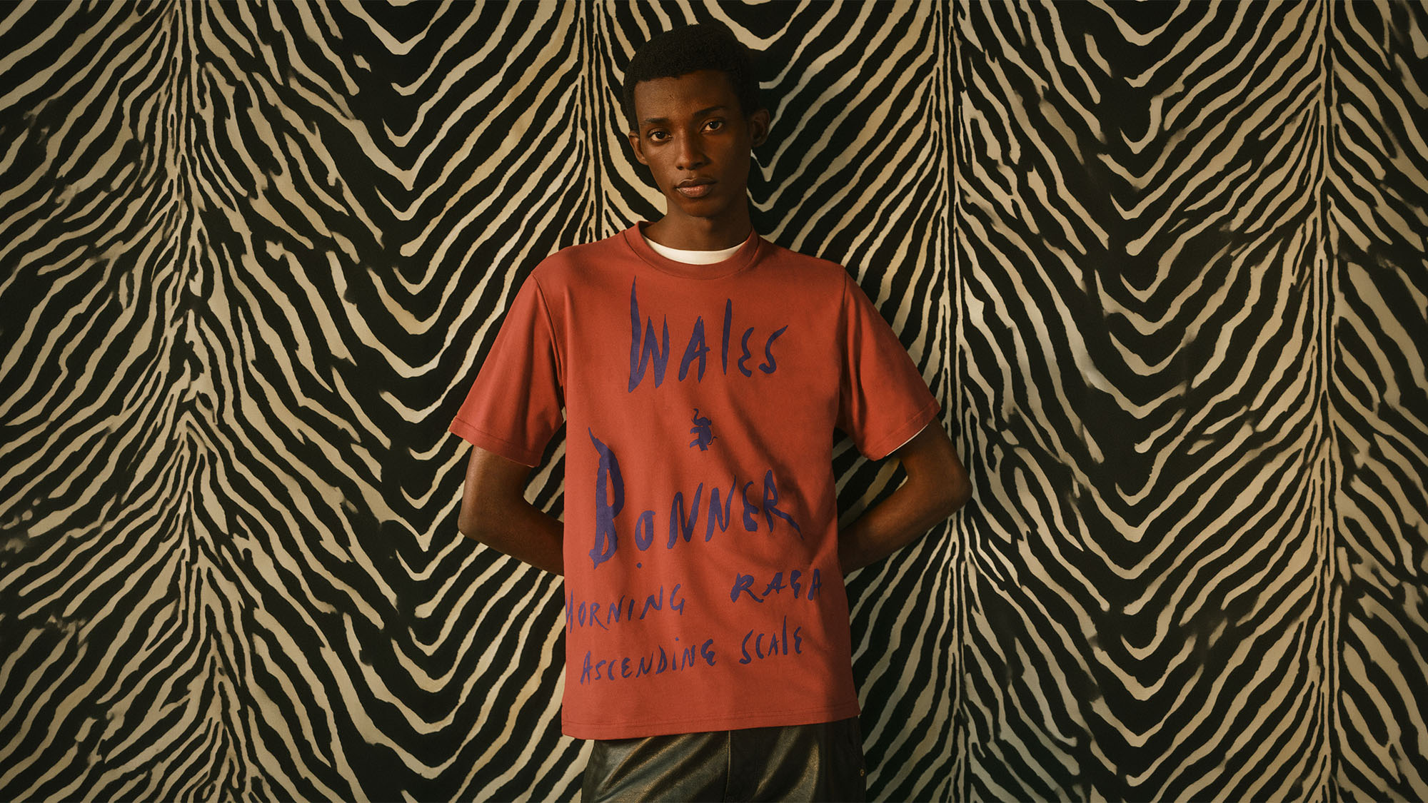Man wearing rust-coloured t-shirt with blue text graphic, standing against black and cream striped background