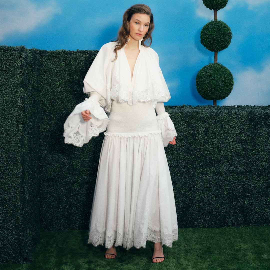 A woman in a white, flowing outfit stands on grass, with a backdrop of greenery and a blue sky with clouds.