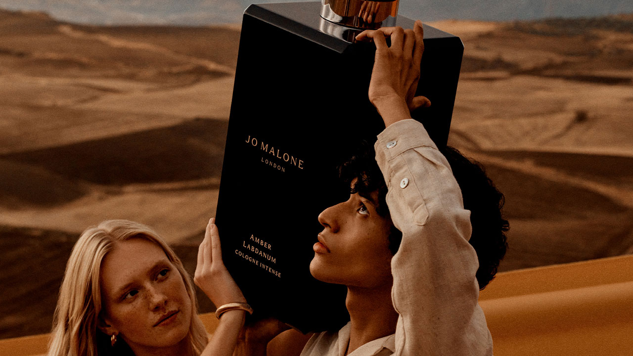 Two people hold a giant Jo Malone perfume bottle in a desert landscape.