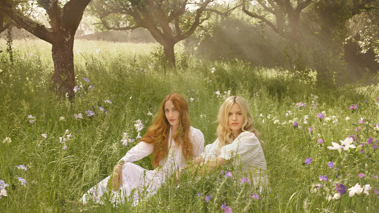 Two women in white dresses sit in a sunlit meadow surrounded by wildflowers and trees.