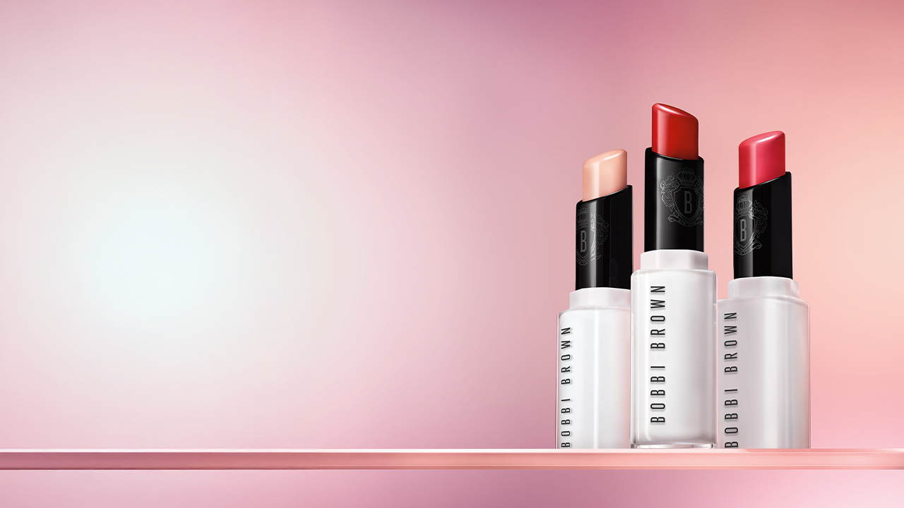 Three Bobbi Brown lipsticks in different shades against a pink gradient background.