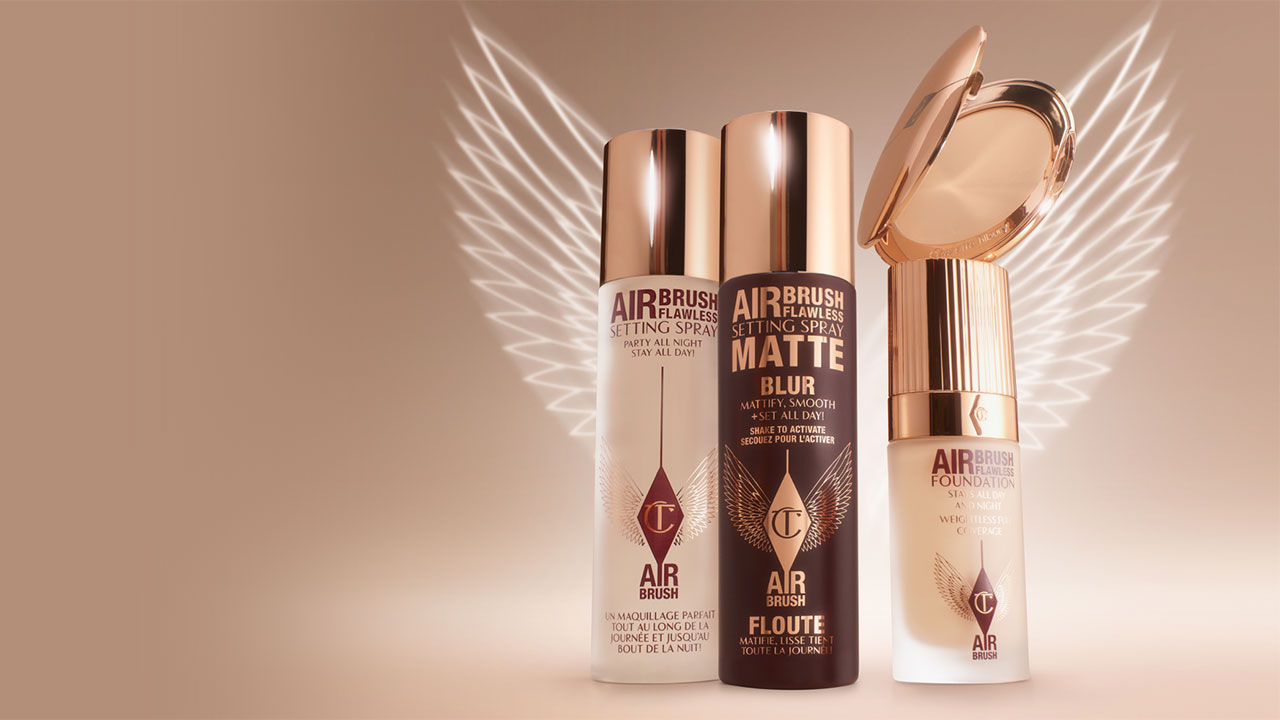 Three makeup products with metallic caps are displayed with white wings in the background on a beige backdrop.