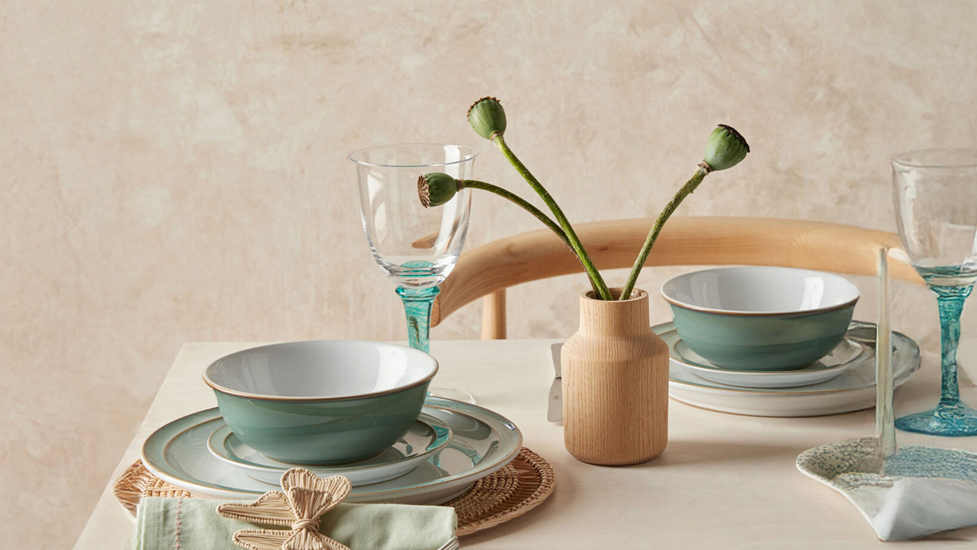 Elegant table setting with green dishes, clear glasses, and a small vase holding buds. Subtle, earthy tones create a minimalist look.