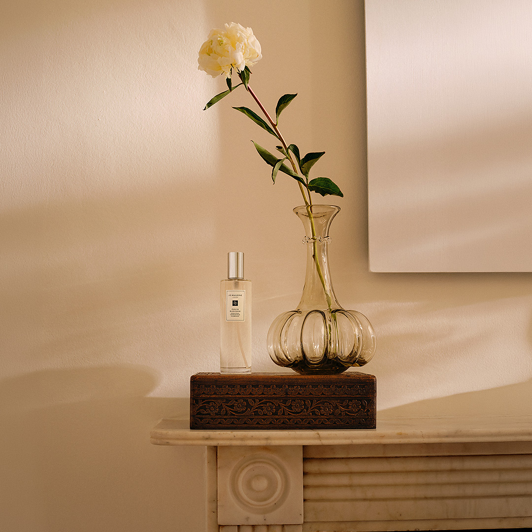 A single flower in a glass vase and a perfume bottle are placed on an ornate box atop a mantelpiece in soft lighting.
