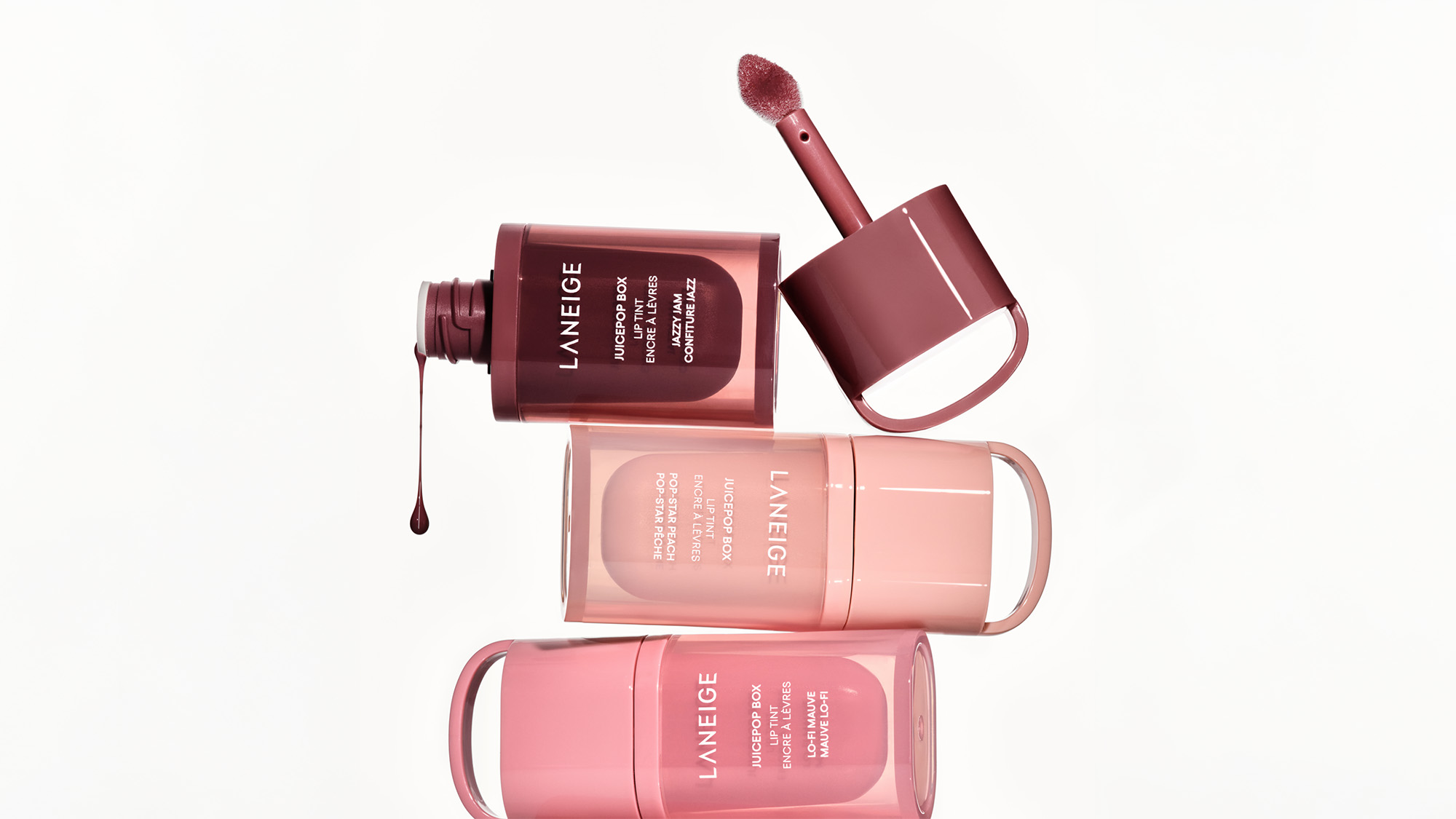 Three Laneige lip products in pink and burgundy shades are displayed creatively with a lip tint applicator.