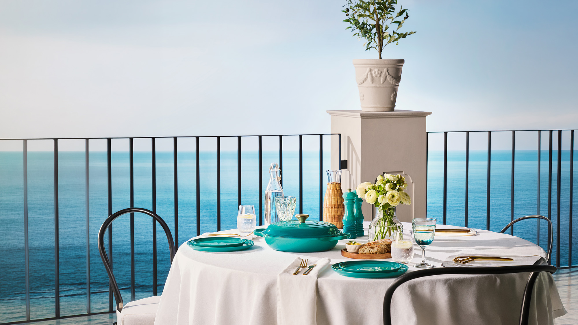 A table set for dining overlooks the ocean, featuring turquoise dinnerware, a white tablecloth, and a potted plant on a ledge.