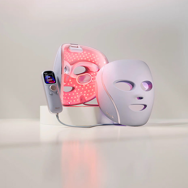 A pair of LED face masks, one white and one pink, with a digital remote control on a white surface.