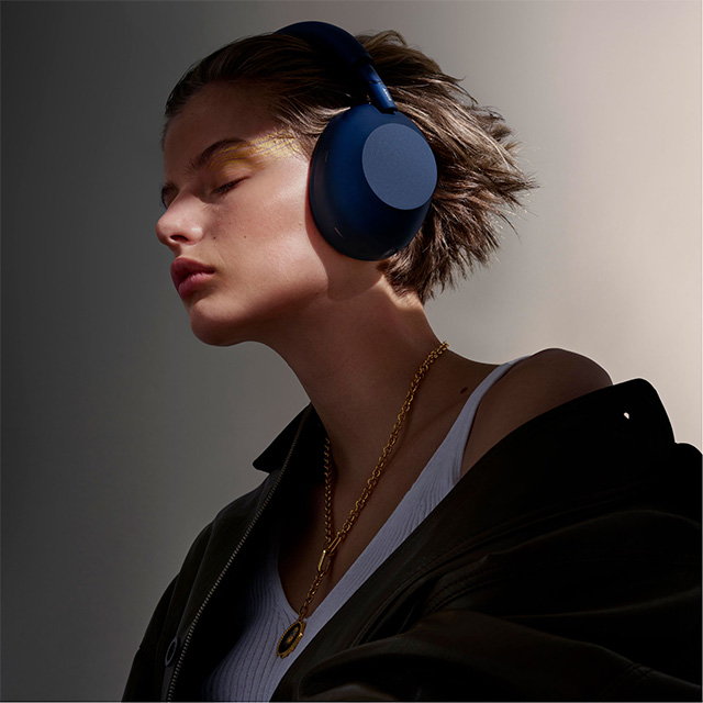 Person with short hair wearing large blue headphones and layered necklaces, eyes closed in soft lighting.