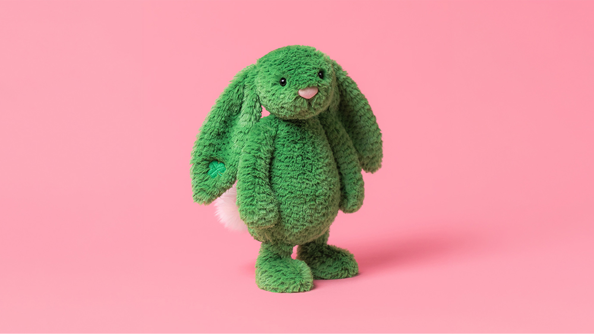 A plush green bunny with long ears and a white tail stands against a pink background.