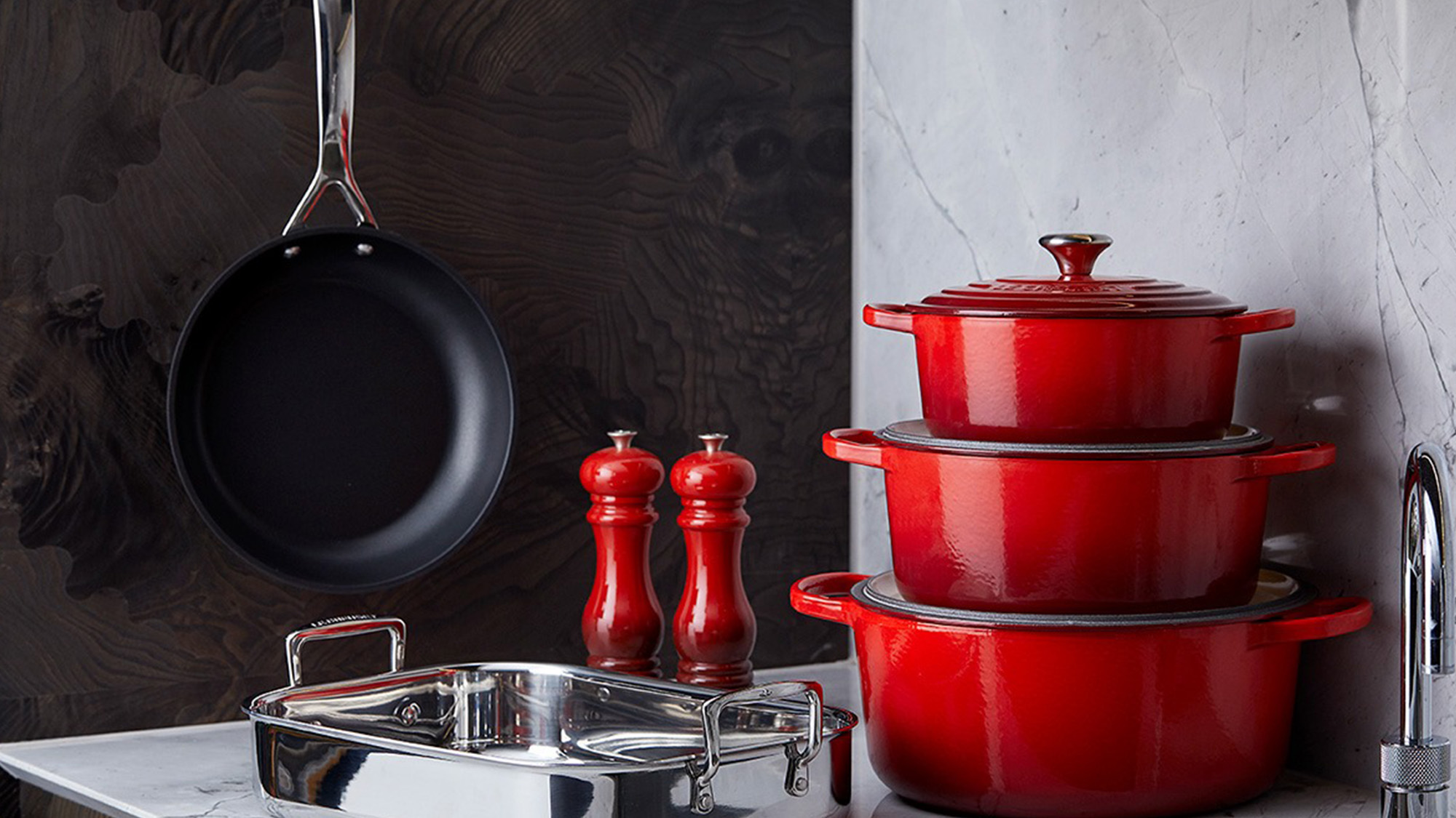 Red cookware set with a frying pan, stacked pots, and red spice grinders on a marble countertop.