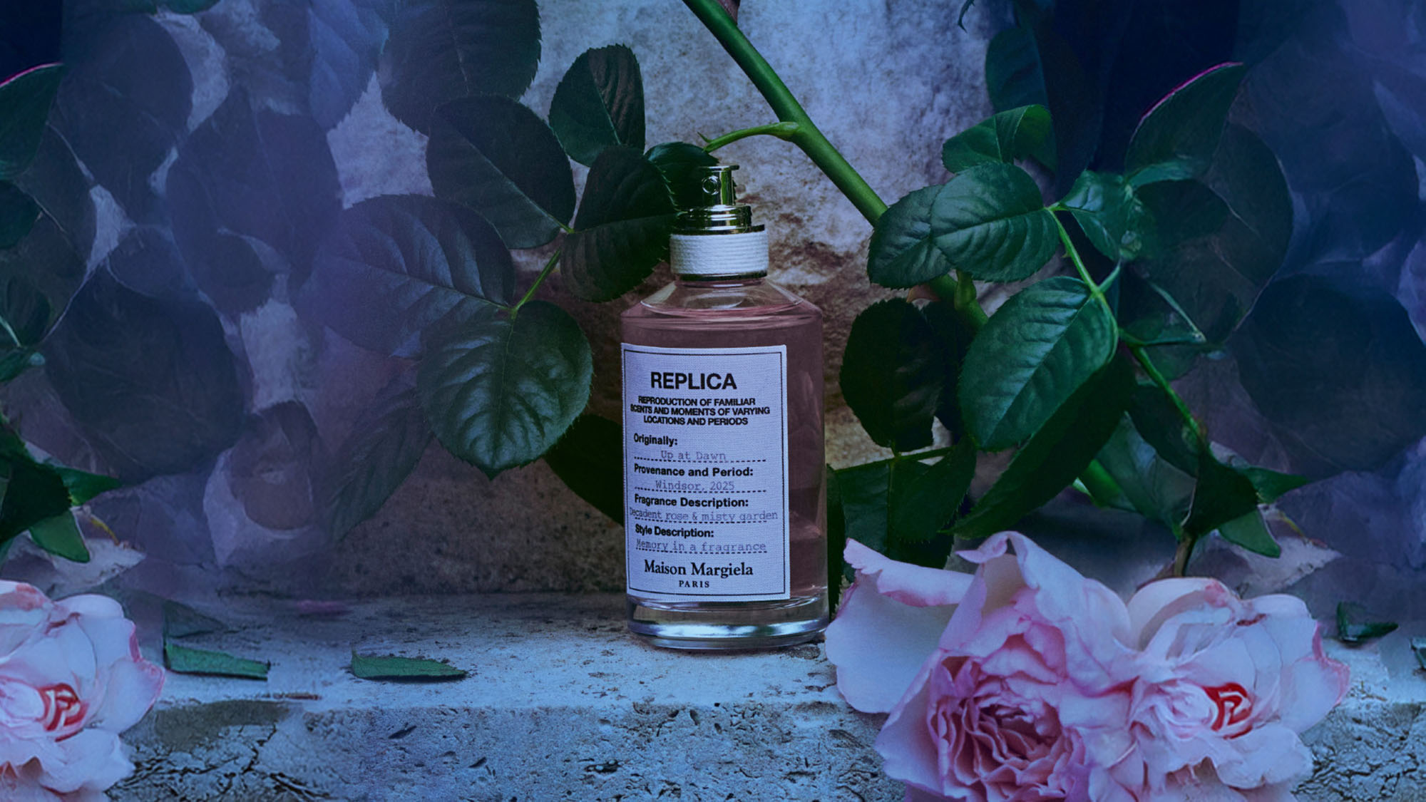 A Maison Margiela 'Replica' perfume bottle is surrounded by roses and green leaves on a textured surface.