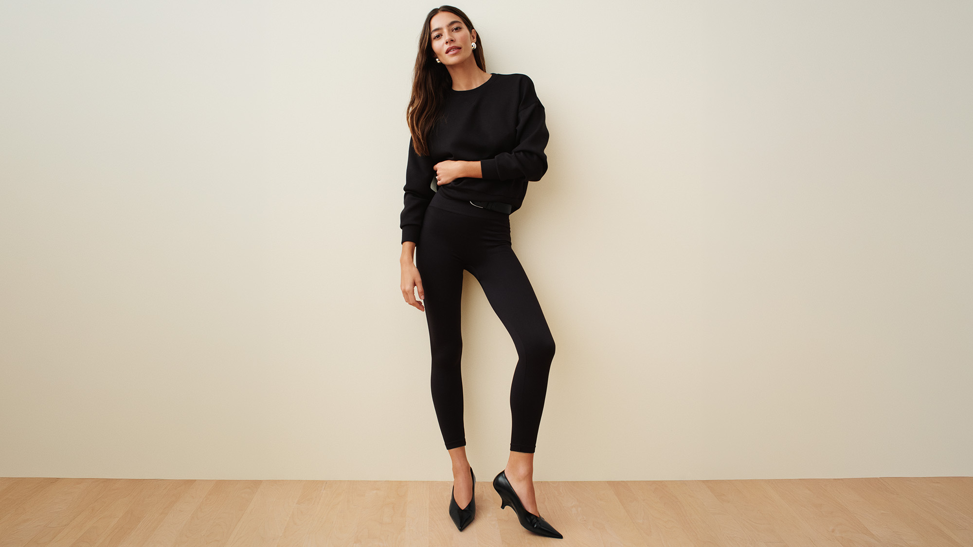 A person in a black outfit, including Spanx leggings, stands against a light wall on a wooden floor.