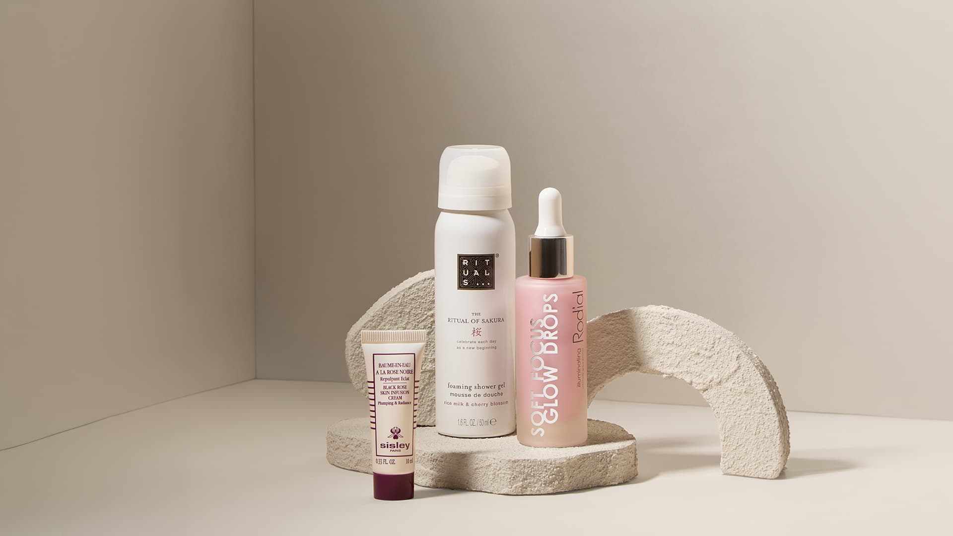 Three skincare products are displayed on a textured, arched beige platform against a neutral backdrop.