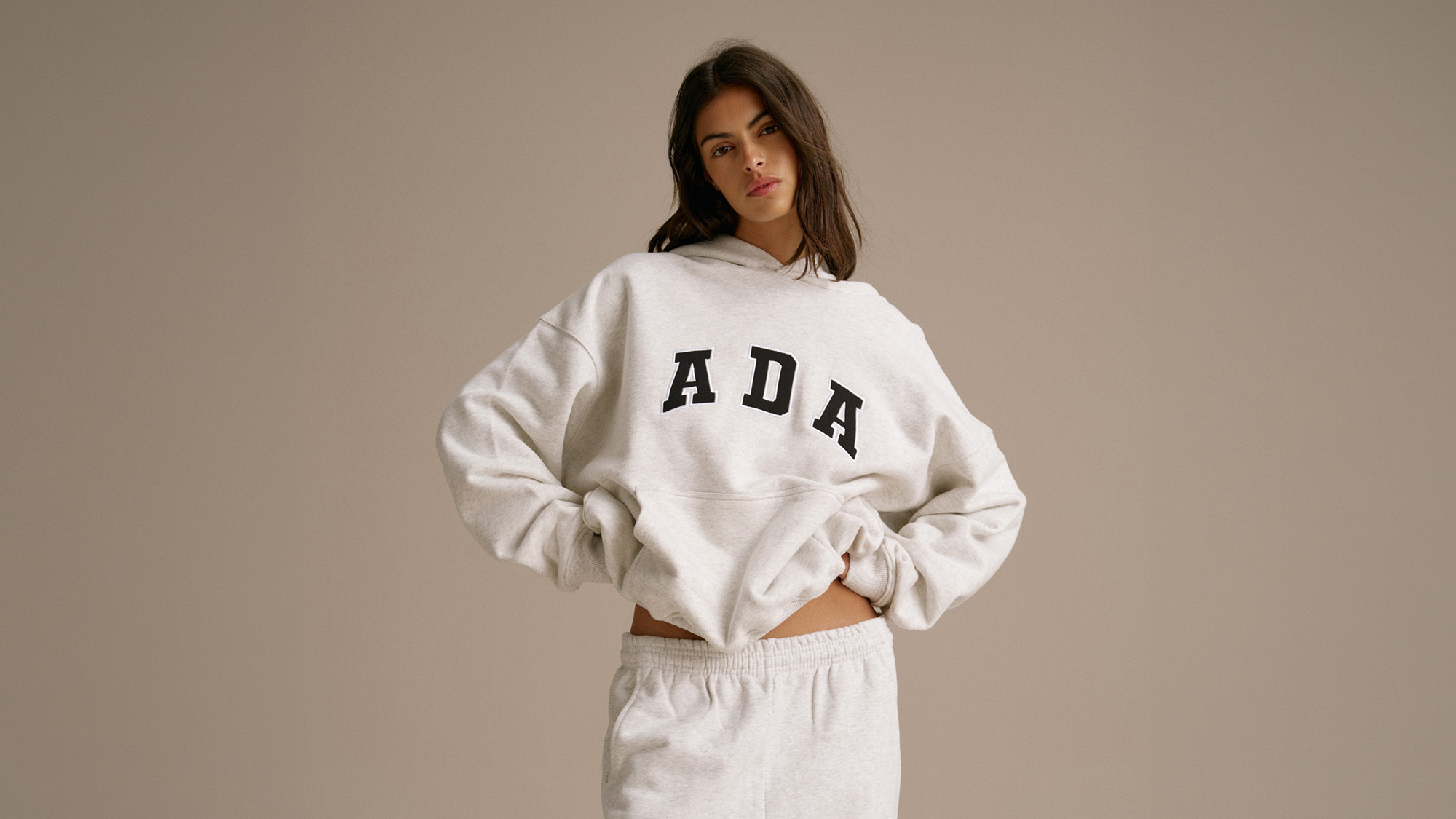 A person wearing a grey hoodie with 'ADA' printed on it and matching sweatpants poses against a neutral background.