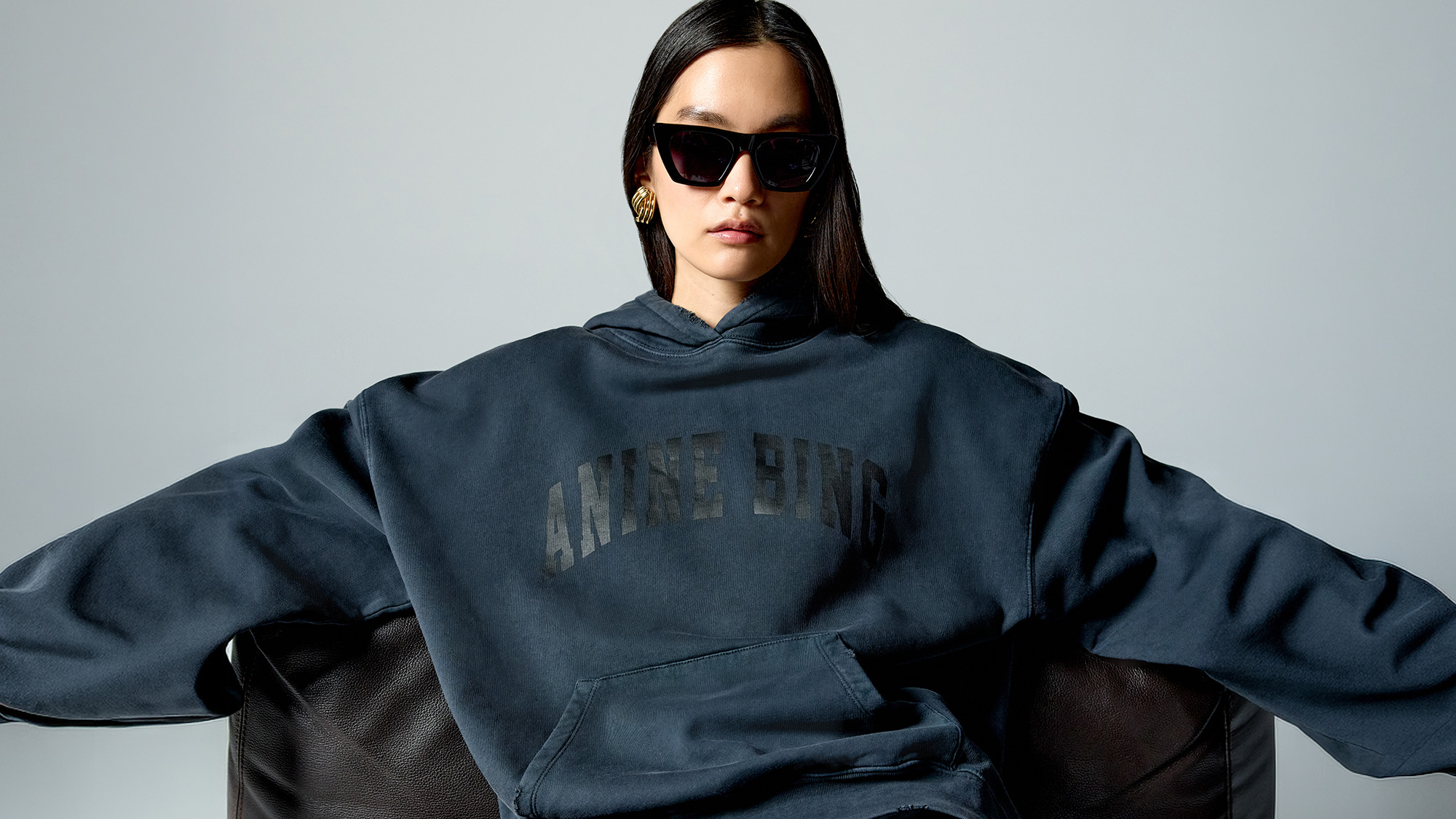 A person in a dark hoodie and leather pants sits on a sofa, wearing sunglasses and gold earrings.