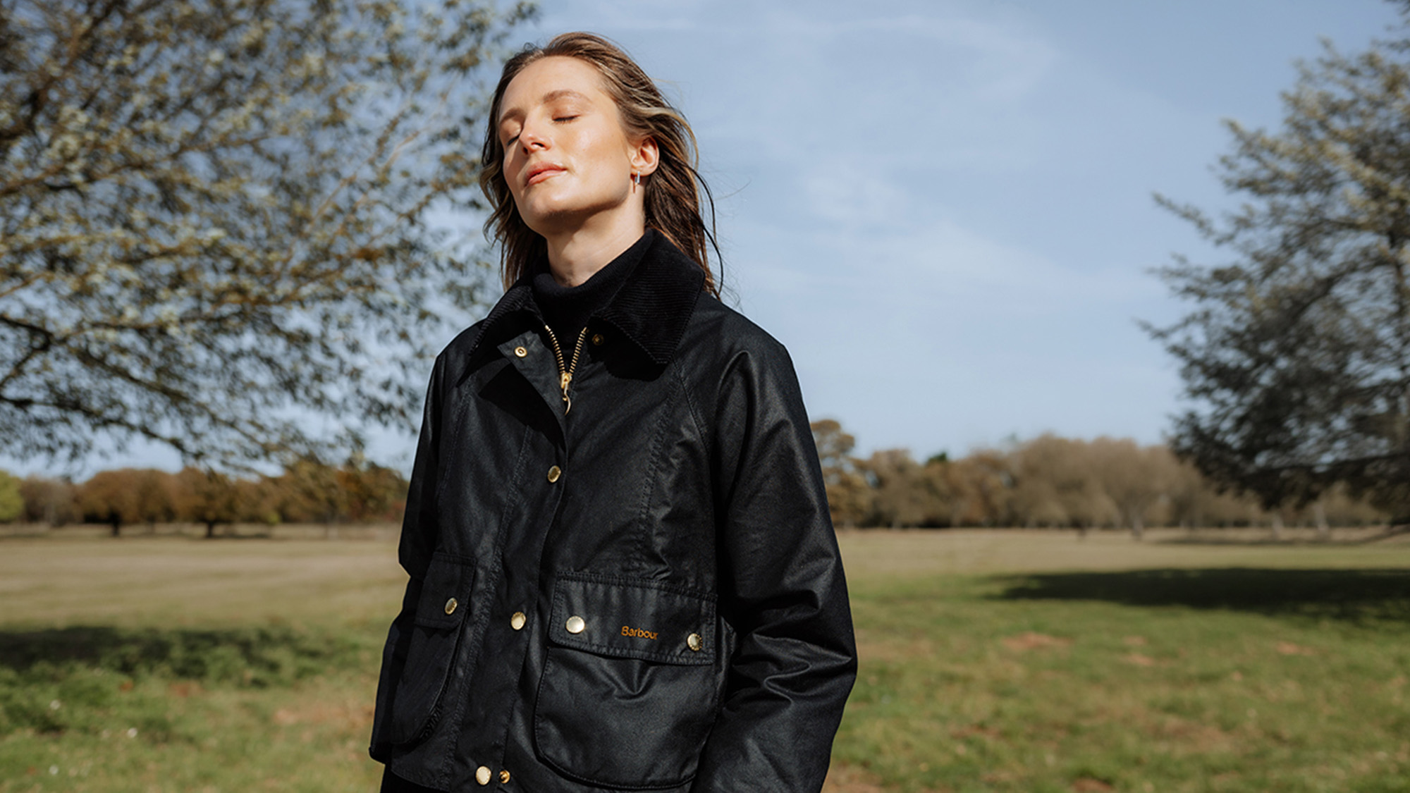 Person wearing a black jacket stands outdoors with eyes closed, enjoying the sunlight.
