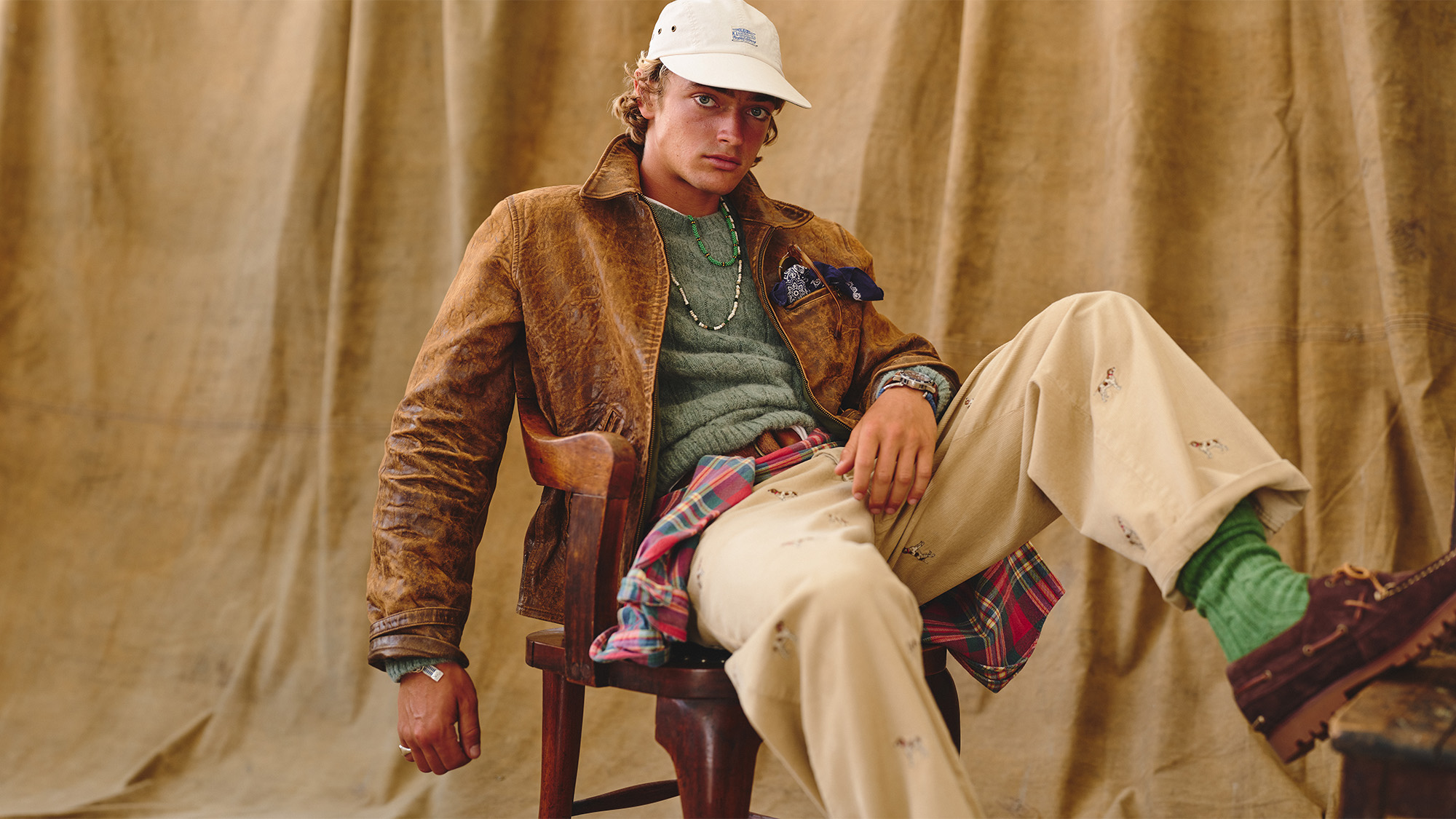 A person wearing a brown jacket, green sweater, white pants with patches, and a white cap, sits on a chair in front of a brown backdrop.