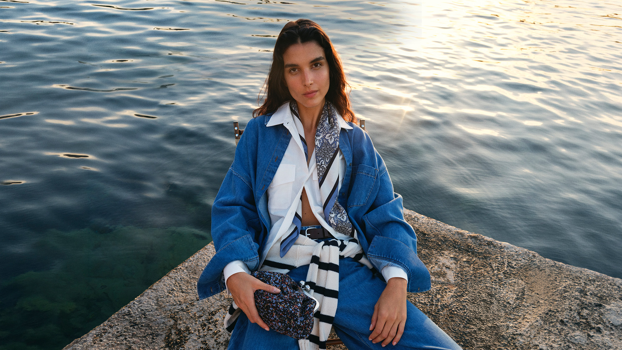 A person in a denim outfit sits on a rock by the water, holding a patterned bag, with a striped scarf draped around their neck.