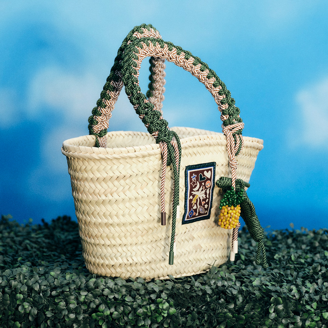 A woman in a dark dress holds a large, brown bag, standing by a rustic window frame.