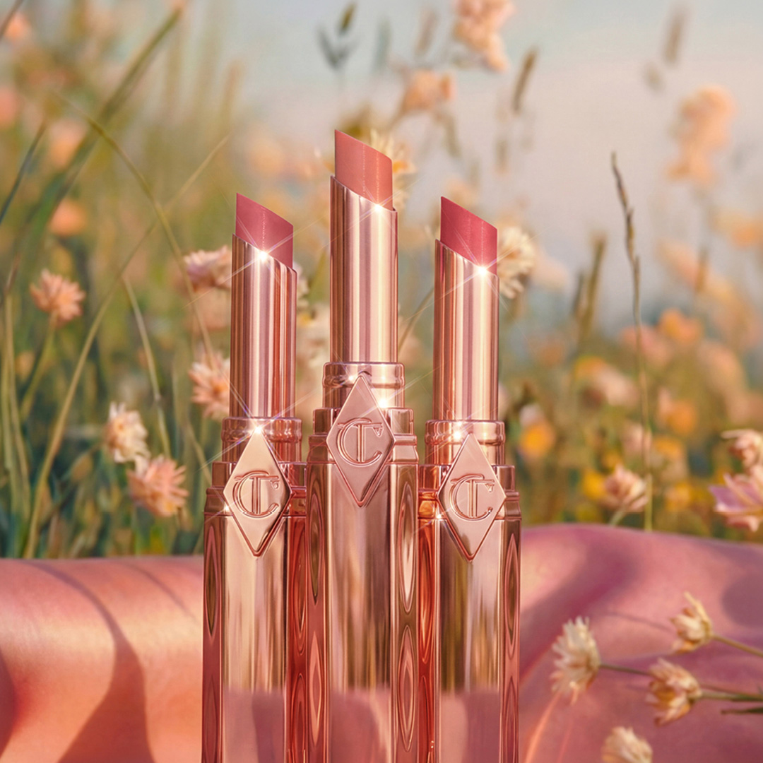 Three rose-gold lipsticks standing on a pink cloth amidst a field of wildflowers.