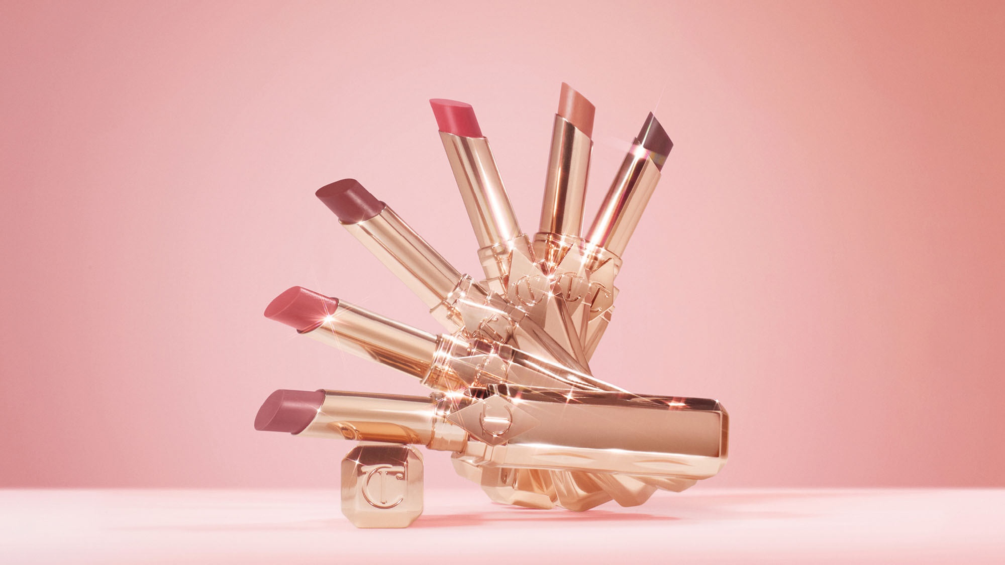 A display of several lipsticks in gold cases, arranged in a fan shape against a pink background.