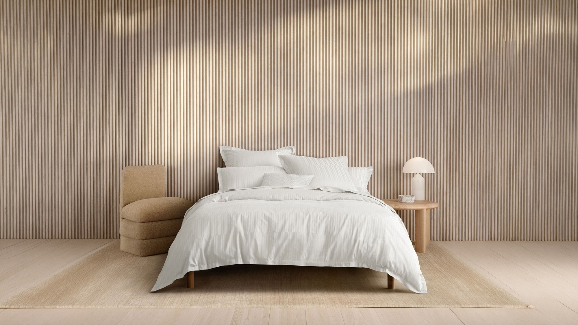 Minimalist bedroom with a white bed, neutral armchair, and vertical line-patterned wall. A small table with a lamp is beside the bed.